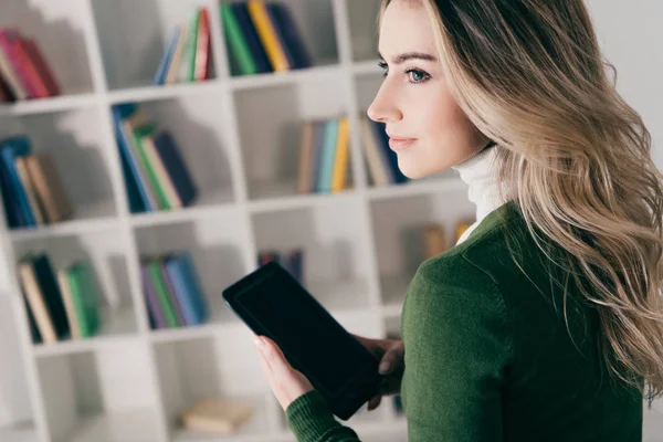 Beautiful woman holding e-book near bookshelf at home — Stock Photo