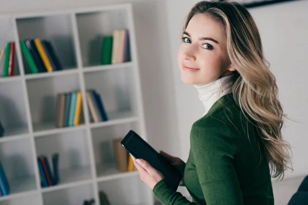 Mulher alegre segurando e-reader e sorrindo perto de estante — Fotografia de Stock