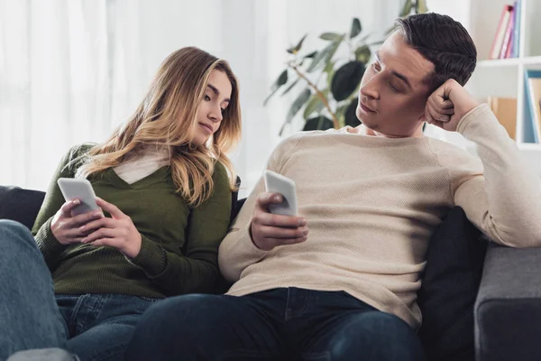 Attrayant femme regardant smartphone dans les mains de petit ami tout en étant assis sur le canapé à la maison — Photo de stock