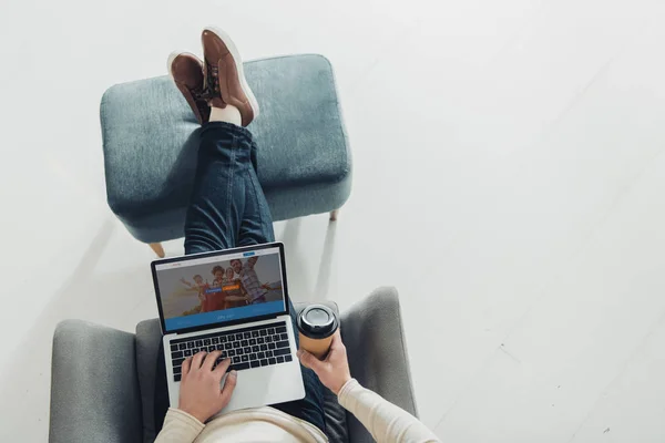 Top view of man holding paper cup and using laptop with couchsurfing website on screen — Stock Photo