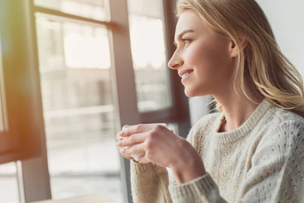 Atractiva mujer sosteniendo taza de té y mirando a la ventana - foto de stock