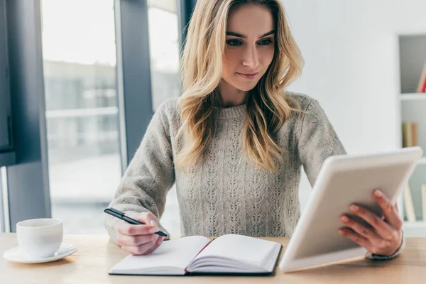 Mulher atraente estudando com tablet digital enquanto segurando caneta perto notebook — Fotografia de Stock