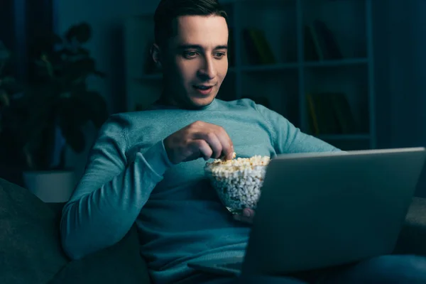 Красивый мужчина смотрит кино на ноутбуке и держит чашу с попкорном дома — стоковое фото