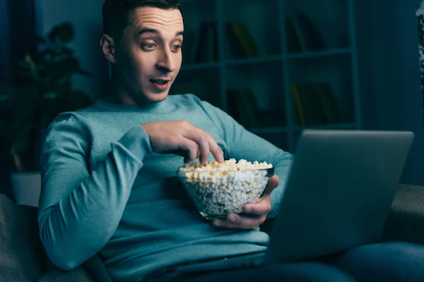 Uomo sorpreso guardando film sul computer portatile e tenendo ciotola con popcorn a casa — Foto stock