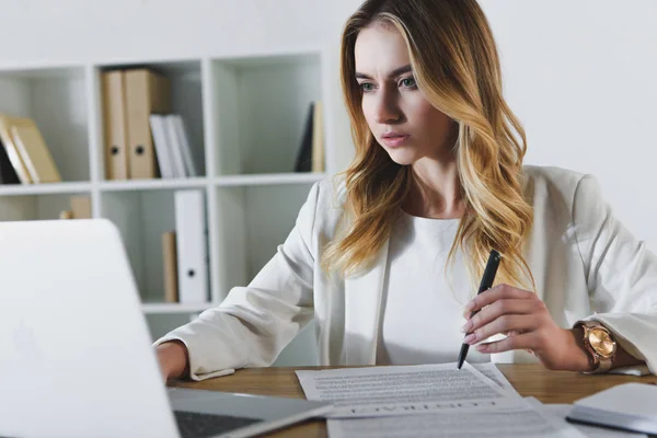 Donna attraente che guarda il computer portatile mentre tiene penna vicino al contratto — Foto stock