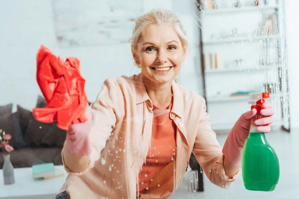 Smiling senior woman cleaning window with rag and spray — Stock Photo