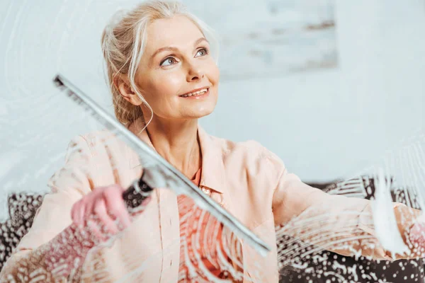 Мрійлива старша жінка прибирає вікно зі скляним склоочисником — стокове фото