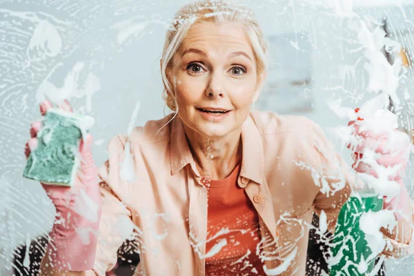 Ventana de limpieza de mujer mayor sorprendida con spray y esponja - foto de stock