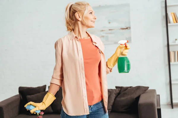 Hermosa mujer mayor en guantes de goma amarilla sosteniendo aerosol y esponja - foto de stock