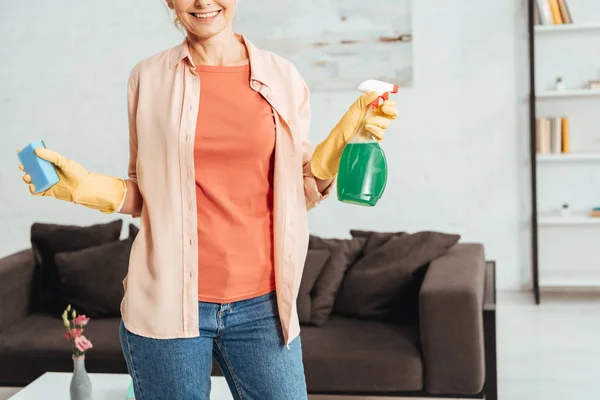 Vista parcial de la mujer en guantes de goma amarilla sosteniendo aerosol de limpieza y esponja - foto de stock