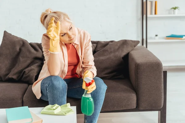 Tired senior woman in rubber gloves sitting on sofa and holding cleaning spray — Stock Photo
