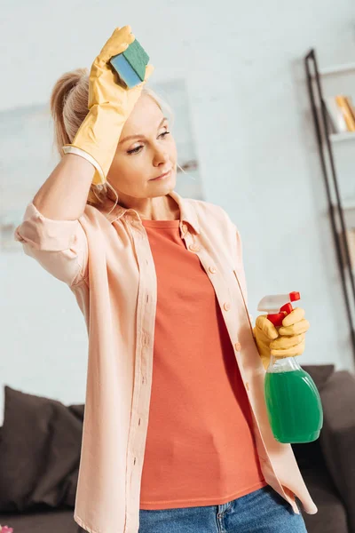 Exhausted senior woman in yellow gloves holding cleaning spray and sponge — Stock Photo