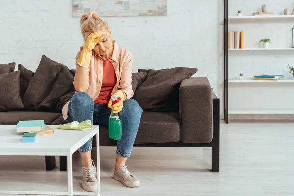 Exhausted senior woman with cleaning spray sitting on sofa — Stock Photo