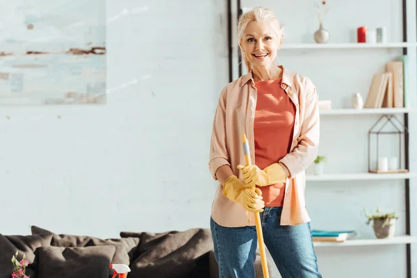 Excited senior woman in yellow gloves posing with mop — Stock Photo