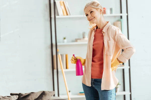 Smiling senior woman in yellow gloves holding spray bottle — Stock Photo