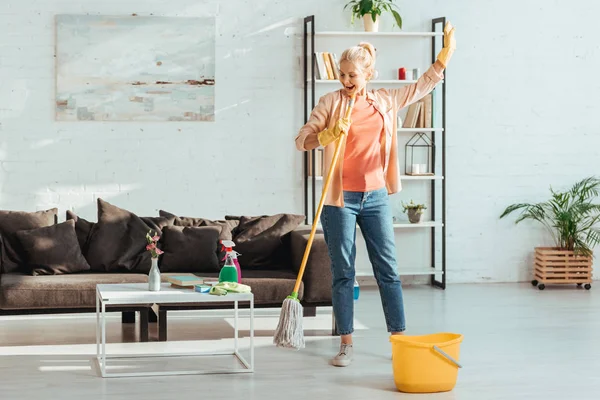 Повний вигляд збудженої старшої жінки прибирання підлоги у вітальні — стокове фото
