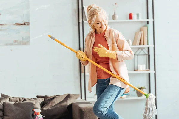 Engraçado mulher sênior tocando esfregona guitarra enquanto limpando casa — Fotografia de Stock