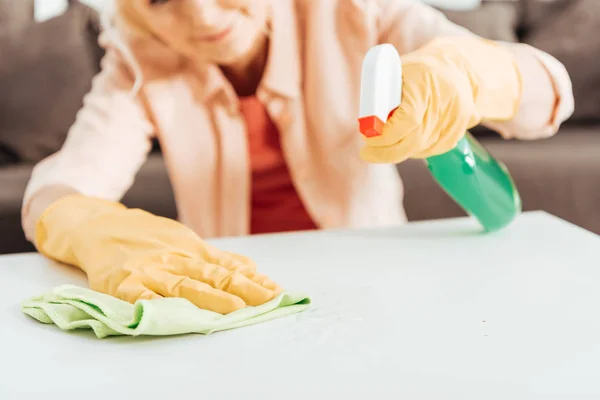 Vue partielle de la femme en gants de caoutchouc jaune nettoyer la surface de la table avec chiffon et pulvérisation — Photo de stock