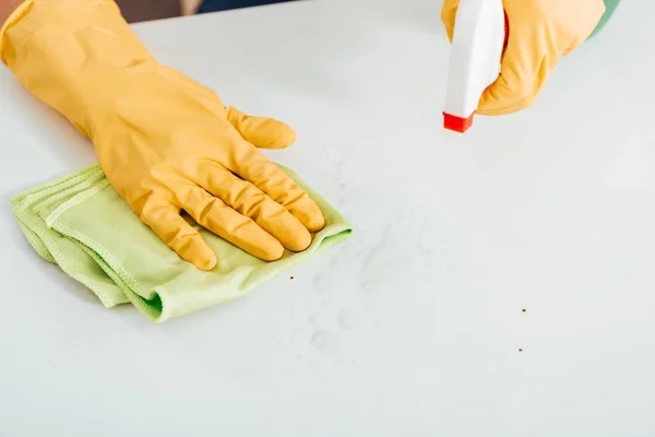 Vista recortada de la mujer en guantes de goma amarilla limpiando la superficie blanca con trapo y spray - foto de stock