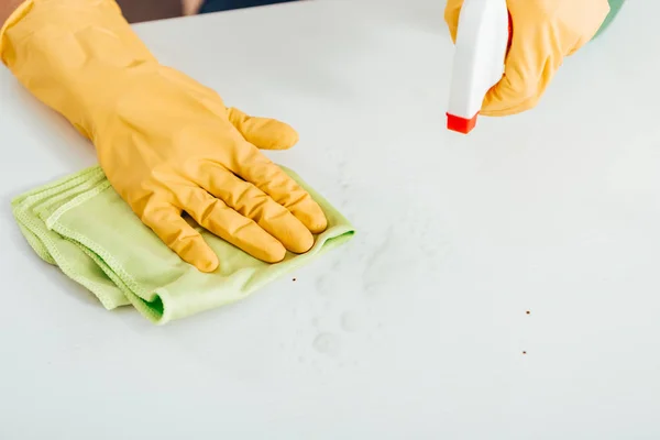 Ausgeschnittene Ansicht einer Frau in gelben Gummihandschuhen, die weiße Oberfläche mit Lappen und Spray säubert — Stockfoto