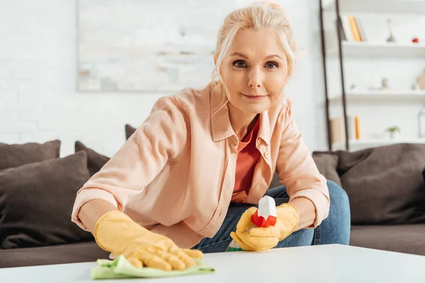 Joyeuse femme âgée en gants de caoutchouc nettoyer la surface de la table avec chiffon et pulvérisation — Photo de stock