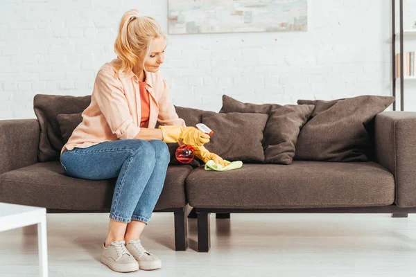 Seniorin in Jeans putzt Sofa mit Spray und Lappen — Stockfoto