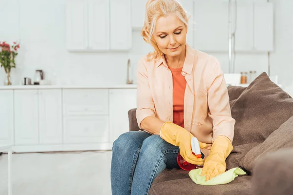 Donna anziana in guanti di gomma divano di pulizia con spray e straccio — Foto stock