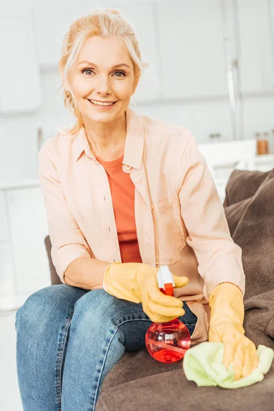 Сміється старша жінка в гумових рукавичках чистить диван з спреєм і ганчіркою — стокове фото
