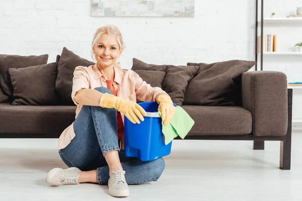 Laughing senior woman sitting on floor and holding bucket with rag — Stock Photo