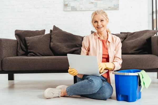 Riendo mujer mayor en guantes de goma usando el ordenador portátil mientras se limpia la casa - foto de stock