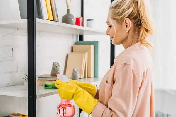 Mujer rubia en guantes de goma amarilla limpiando estantes de libros - foto de stock