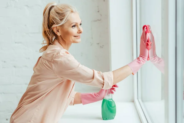 Mulher sênior sorridente em luvas rosa janela de limpeza com pano e spray — Fotografia de Stock