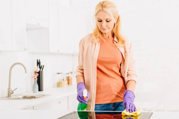 Loira sênior mulher lavar cozinha fogão com pano e spray — Fotografia de Stock