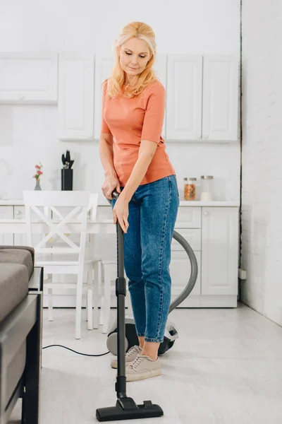 Старша жінка в джинсах прибирає підлогу з пилососом — стокове фото