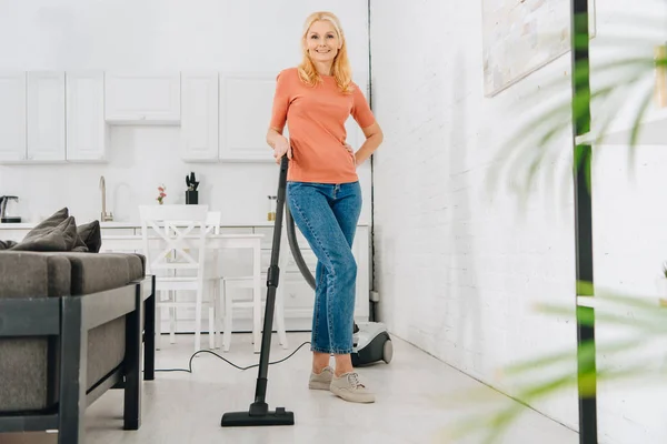 Mulher sênior feliz em jeans casa de limpeza com aspirador de pó — Fotografia de Stock