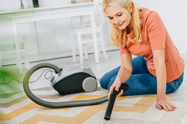 Smiling blonde woman sitting on carpet and using vacuum cleaner — Stock Photo