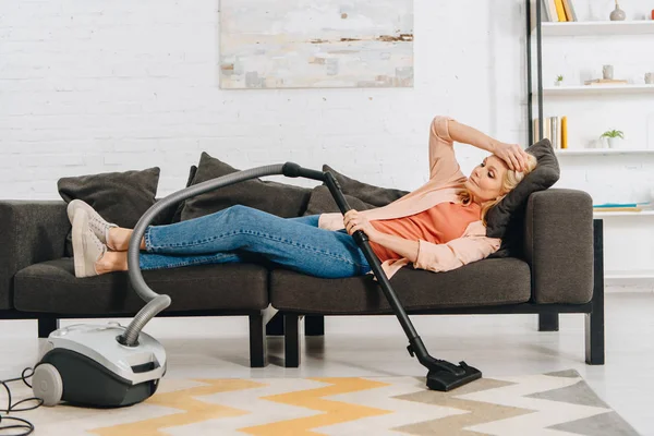 Erschöpfte Frau mit Staubsauger auf Sofa liegend und Kopf berührt — Stockfoto