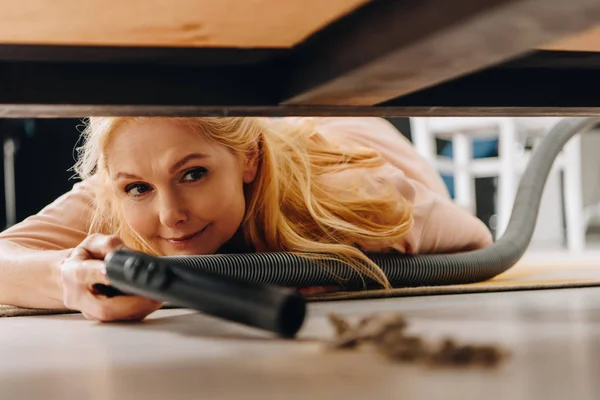 Smiling senior woman using vacuum cleaner under furniture — Stock Photo