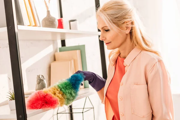 Senior woman in rubber glove cleaning shelves with bright duster — Stock Photo