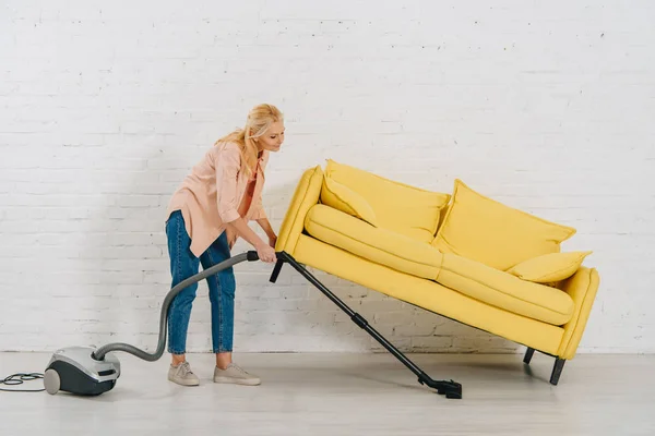 Ansicht der Seniorin mit Staubsaugerboden unter gelbem Sofa in voller Länge — Stockfoto