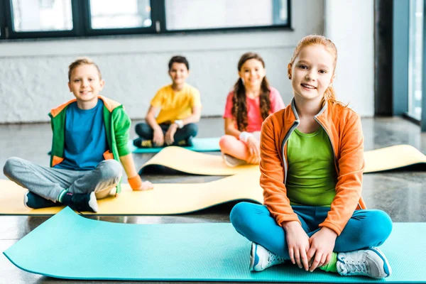 Niños sentados en colchonetas de fitness y mirando a la cámara con sonrisa - foto de stock