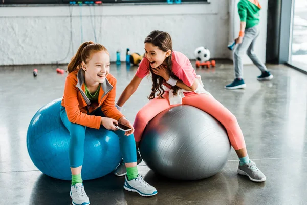 Lachende Kinder mit Smartphone sitzen auf Fitnessbällen — Stockfoto