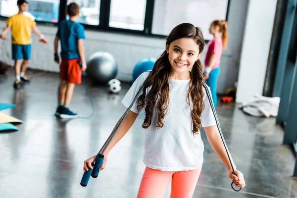 Cute brunette kid holding jump rope and smiling at camera — Stock Photo