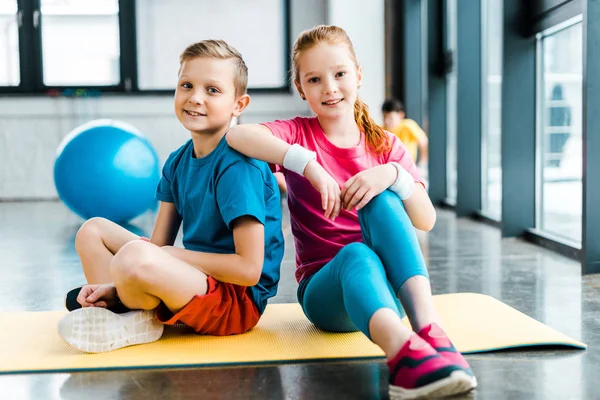 Amigos preadolescentes sentados en una colchoneta de fitness en el gimnasio - foto de stock