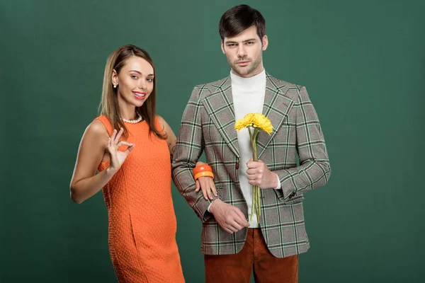 Bella donna facendo ok segno mentre l'uomo in abiti vintage tenendo fiori isolati su verde — Foto stock