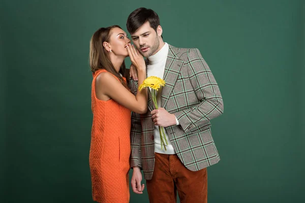 Beautiful woman in vintage clothes whispering something to boyfriend with flowers isolated on green — Stock Photo