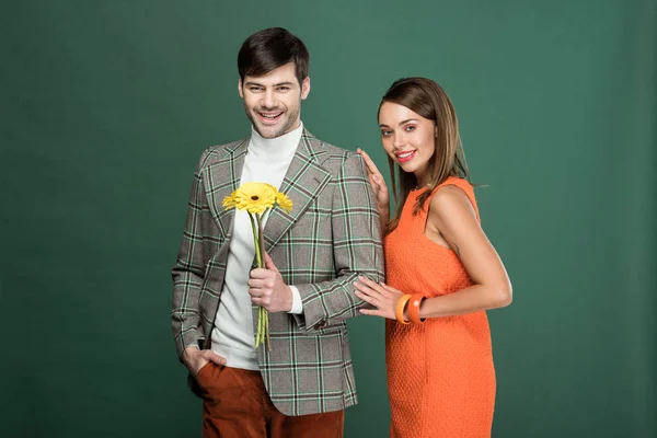 Schöne Frau blickt in die Kamera und umarmt Mann in Vintage-Kleidung mit Blumen isoliert auf grün — Stockfoto