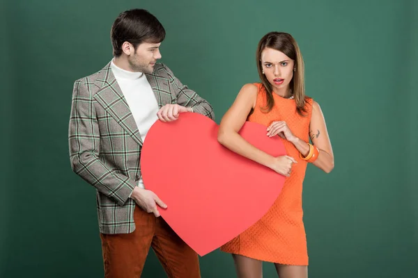 Мужчина смотрит на недовольную женщину, держащую бумажную карточку в форме сердца, изолированную на зеленом — стоковое фото