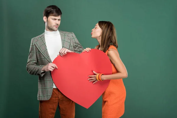 Hombre insatisfecho mirando a la hermosa mujer haciendo pucheros labios y sosteniendo tarjeta de papel en forma de corazón aislado en verde - foto de stock