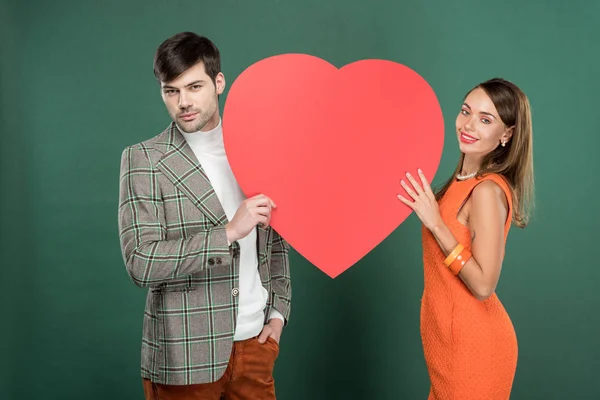 Hermosa pareja sosteniendo la tarjeta de papel en forma de corazón y mirando a la cámara aislada en verde - foto de stock